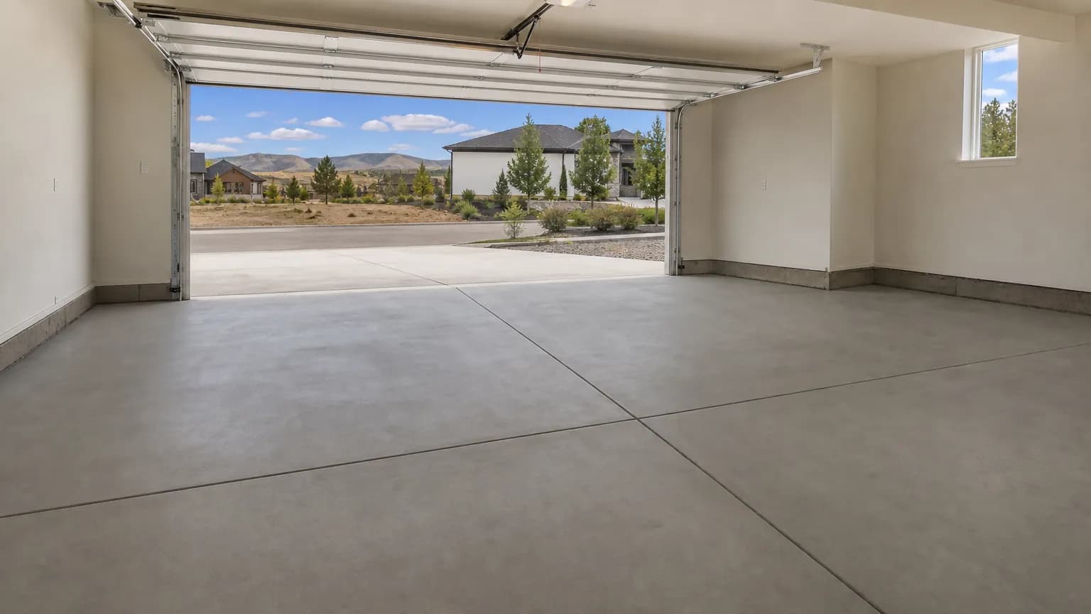 Empty two-car garage with a smooth, freshly troweled gray concrete floor and a saw-cut joint, with the door open to daylight