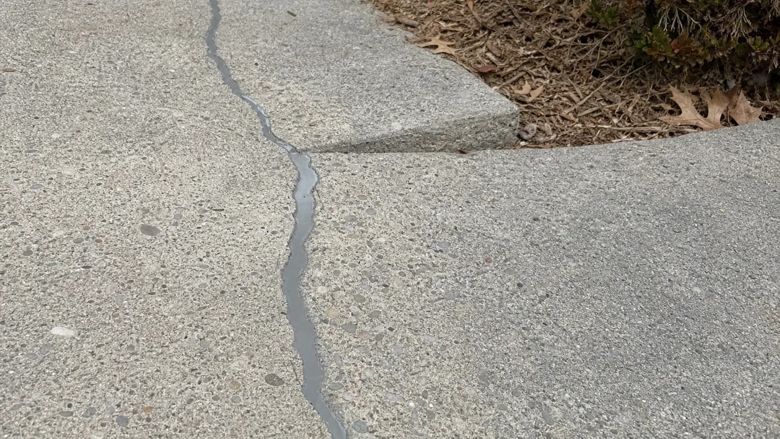 Older concrete sidewalk slab with a long crack routed and filled with gray sealant next to a slightly sunken panel