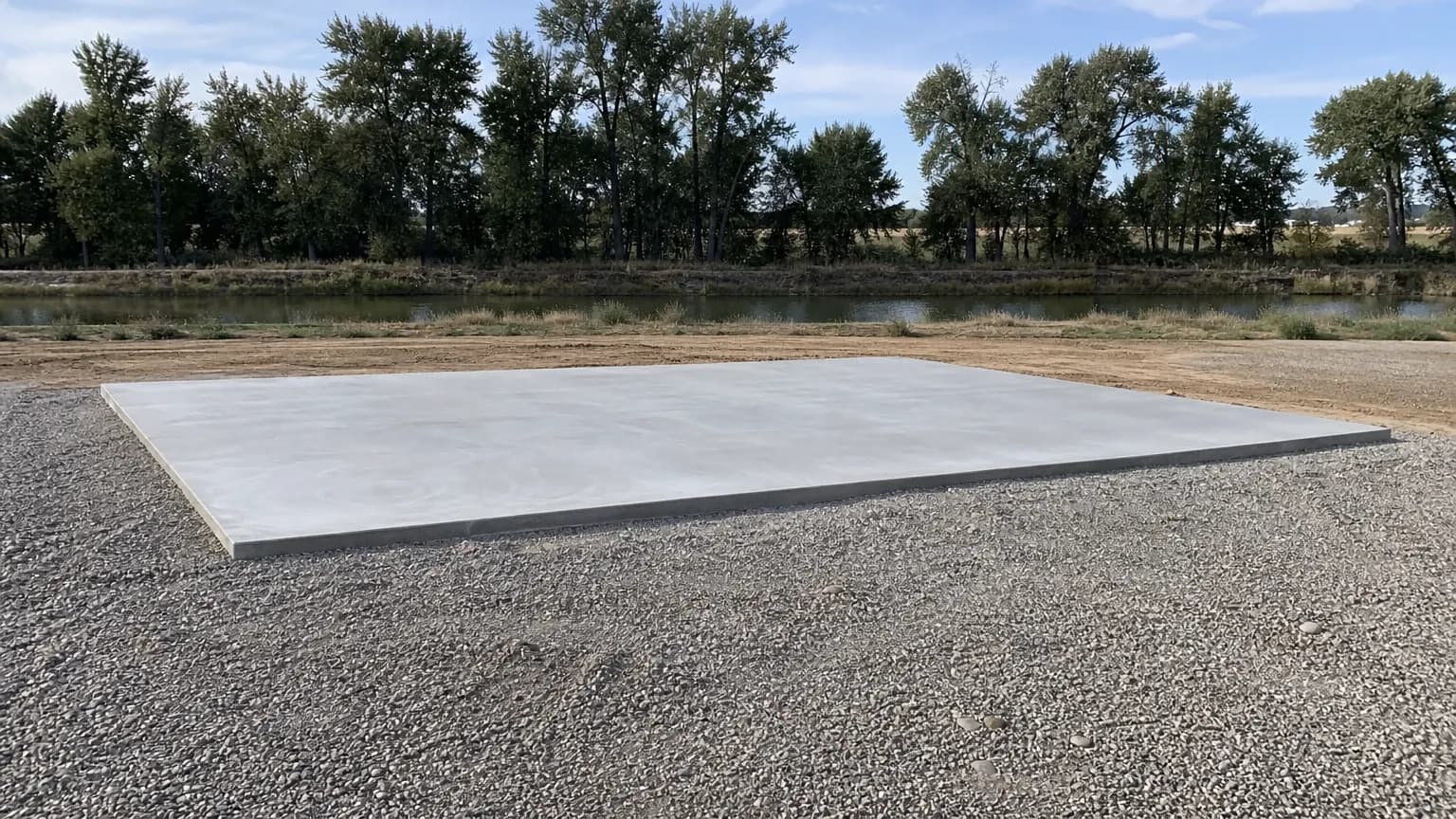 Plain cured concrete slab pad on a gravel apron on a semi-rural lot, with cottonwoods along the water behind